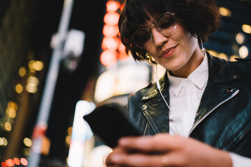 Wall Mural - Below view of attractive hipster girl dressed in leather jacket choosing song in playlist downloaded on smartphone for listening in earphones while walking in night downtown with illumination lights