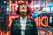 Stylish Brunette Woman In Trendy Apparel And Eyewear Looking Up Enjoying Nightlife In City.Gorgeous Hipster Girl Dressed In Leather Jacket Standing Outdoors On Street With Neon City Illumination