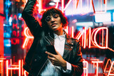 Fototapeta  - Beautiful brunette woman in trendy apparel and eyewear enjoying nightlife in city listening music in earphones and moving to sound, gorgeous hipster girl dancing outdoors on neon city illumination .