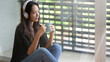 © bongkarn - Relaxed woman listening to music with headphones and holding a coffee cup while sitting on floor at bedroom