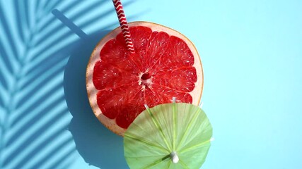 Wall Mural - Grapefruit (orange) rotating with a cocktail umbrella in the sunlight on a colored blue background. Fruit juices, relaxation, tropics and relaxation concept.