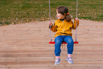 Wall Mural - Happy little girl swinging on a swing in the Park in beautiful autumn, or cold summer. Cute child 3-4 years in a jacket and jeans having fun on a playground outdoors. Adorable girl in a park.