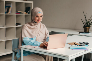 young pretty modern muslim woman in hijab working on laptop in office room, education online, remote work freelancer, typing thinking concentrated