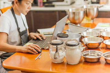 Female baker working on laptop with bakery ingredient on table.