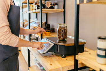 Wall Mural - Female shop owner weighs glass jar with dark brown whole grains of pasta. Woman assistant in zero waste shop. Weighing dry goods in plastic free grocery store. Sustainable shopping at local business