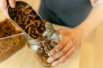 Wall Mural - Dark brown whole grains of pasta are poured in glass jar. Close up of unrecognizable woman is pouring pasta to a jar from bulk dry food dispensers at zero waste shop, defocused.