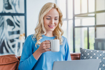 Wall Mural - Young woman working on laptop computer while sitting on sofa at home with morning cup of coffee