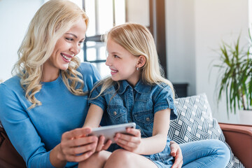 Wall Mural - Beautiful young mom and her little daughter using smartphone and smiling while sitting on sofa at home