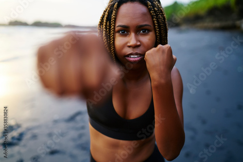 Half length portrait of afro american female boxer doing exercises during workout on seashore while looking at camera.Dark skinned sportwoman in active wear boxing with hands on beach during training