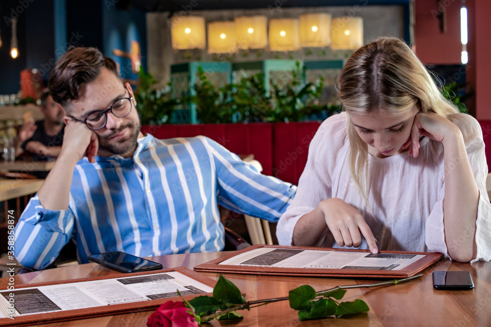 Happy young romantic couple choosing food from menu at dinner in a beautiful fancy restaurant. Man bored, waiting for his hesitant woman to decide. Fine dining concept.