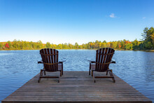 Two Muskoka Chairs Sitting On A Wood Dock Facing A Lake In A Calm Autumn Season Sunny Day, With The Chairs In The Sun Shadow.