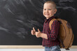 © Andrii Lysenko - A happy schoolboy of 6-7 years old is standing at school near the blackboard with a school bag. He shows the class with two thumbs. September 1. Back to school.