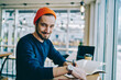© BullRun - Portrait of cheerful caucasian hipster guy checking time for learning in cafe interior, happy male freelancer looking at camera showing wristwatch using time management for complete remote job.