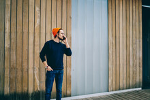 Caucasian Man In Trendy Wear And Orange Hat Standing Near Urban Setting Wall Talking On Mobile Phone, Serious Young Hipster Guy Having Conversation In Romaing On Cellphone Spending Time On Street.