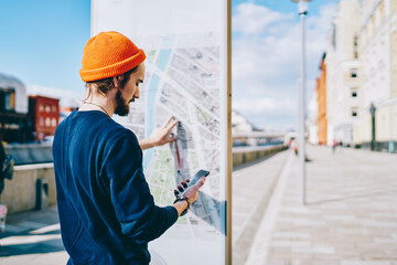 Wall Mural - Hipster guy checking schedule of public transport on modern electronic board with information, back view of male traveler searching location on mobile phone application and on map board on street.