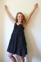 cute little girl with long blond hair smiles, raising her hands up against a white wall