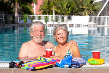 Wall Mural - Close up of an elderly couple of people smiling and enjoying free time in the pool. Sun on the face. Floating glasses with straws. Towels full of colors. Serene retirement days