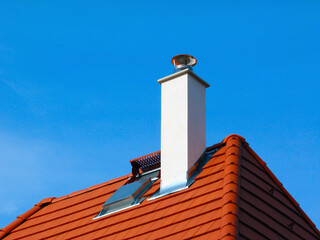 sloped red clay tile roof and ridge detail. new pitched residential roof. service platform. stainless steel smoke stack. white chimney. construction and building materials. blue sky and sunlight.