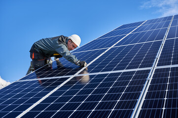 Wall Mural - Beautiful and shiny solar battery surface and man worker wearing a uniform, standing on ladder. Male electrician installing solar modules on sunny day, low angle view. Concept of alternative energy