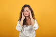 Headache grimacing pain holds the back of neck indicating location. Fatigue during workaholism labor. Young attractive woman, dressed white blouse, with brown eyes, curly hair, yellow background