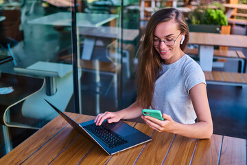 Wall Mural - Happy working woman freelancer in wireless headphones and eyeglasses using phone and laptop for working online in cafe. Modern people with digital smart mobility lifestyle