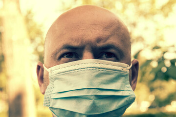 Portrait of young man wearing medical mask posing in sunny park. Close up portrait of a man looking away thoughtfully, wearing medical protective mask outdoors in the park. toned