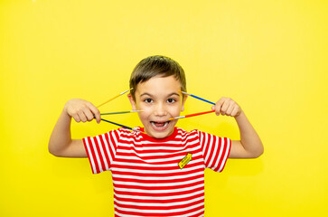 Portrait of emotional happy boy in a striped T-shirt smiles and holds multi-colored paint brushes on a yellow background