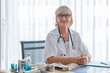 © eakgrungenerd - Female senior doctor wear uniform and spectacles with stethoscope sitting at work table in hospital.