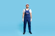 © khosrork - Full length handyman in overalls, cap and protective gloves standing, looking calm and positive at camera. Profession of service industry, house repair. indoor studio shot isolated on blue background