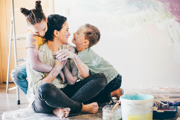 funny children with paints on skin and clothes hug mother sitting on new apartment floor against coloured wall and metal wooden ladders