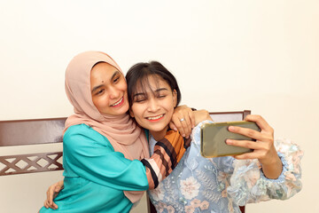 Wall Mural - Two young Asian Malay Muslim woman wearing headscarf at home office student sitting at table phone computer book document selfie self portrait with smartphone