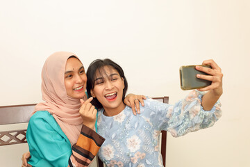 Wall Mural - Two young Asian Malay Muslim woman wearing headscarf at home office student sitting at table phone computer book document selfie self portrait with smartphone care love