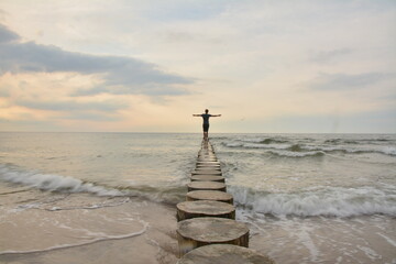 A young boy standing on stilts in the sea with his arms outstretched, looking at the sunet