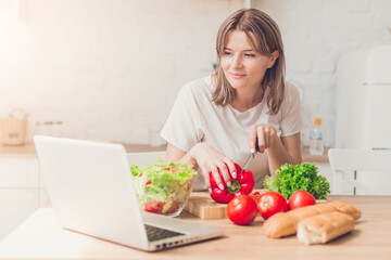 Woman cutting vegetables on salad and view recipes in laptop on kitchen.