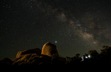 Wall Mural - starry night sky showing milky way core rising above skull rock in Joshua tree national park.  The rock is lit orange by a few hikers' head lamps.  Jupiter is seen rising above the rock's dome. 