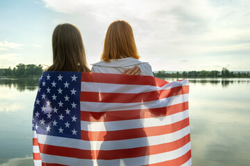 Two young friends women with USA national flag on their shoulders hugging together outdoors on lake shore. Patriotic girls celebrating United States independence day.