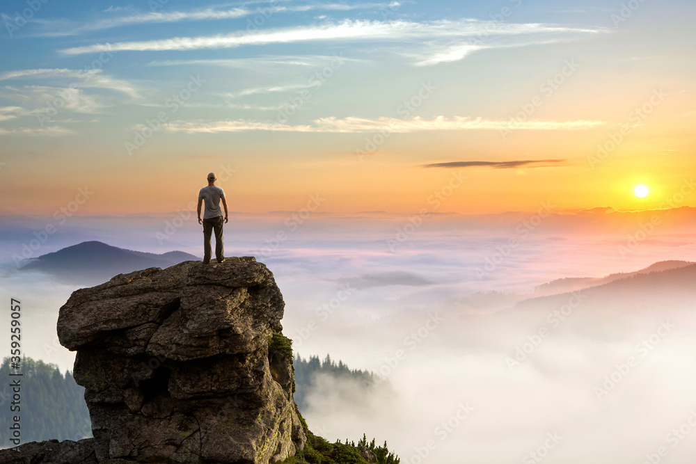 Small silhouette of hiker man enjoying beautiful sunrise in morning mountains. Traveler with raised hands standing on mountain with white fog below. - obrazy, fototapety, plakaty 