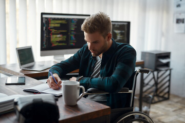 Side view of young concentrated male web developer in a wheelchair making some notes while sitting at his workplace with multiple computer screens on the background
