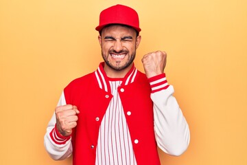 Handsome sporty man with beard wearing baseball jacket and cap over yellow background celebrating surprised and amazed for success with arms raised and eyes closed