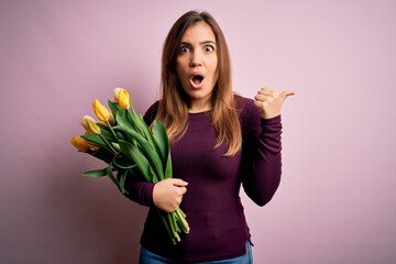 Sticker - Young blonde woman holding romantic bouquet of yellow tulips flowers over pink background Surprised pointing with hand finger to the side, open mouth amazed expression.