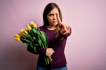 Canvas Print - Young blonde woman holding romantic bouquet of yellow tulips flowers over pink background Pointing with finger up and angry expression, showing no gesture