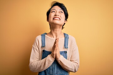 Poster - Young beautiful asian girl wearing casual denim overalls over isolated yellow background begging and praying with hands together with hope expression on face very emotional and worried. Begging.