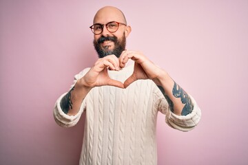 Wall Mural - Handsome bald man with beard and tattoo wearing glasses and sweater over pink background smiling in love doing heart symbol shape with hands. Romantic concept.