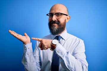 Canvas Print - Handsome business bald man with beard wearing elegant tie and glasses over blue background amazed and smiling to the camera while presenting with hand and pointing with finger.