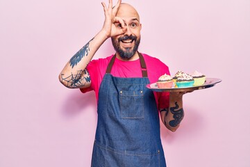 Sticker - Young handsome man wearing apron holding cupcake smiling happy doing ok sign with hand on eye looking through fingers