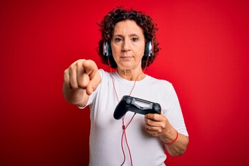 Sticker - Middle age curly hair gamer woman playing video game using joystick and headphones pointing with finger to the camera and to you, hand sign, positive and confident gesture from the front