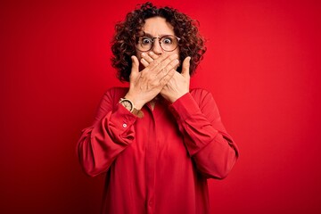 Poster - Middle age beautiful curly hair woman wearing casual shirt and glasses over red background shocked covering mouth with hands for mistake. Secret concept.