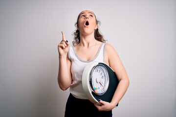 Wall Mural - Young beautiful woman with curly hair holding weighing machine over white background amazed and surprised looking up and pointing with fingers and raised arms.