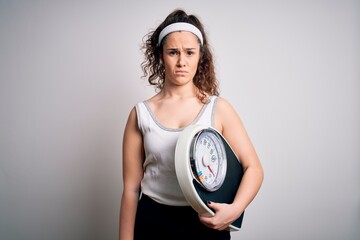 Sticker - Young beautiful woman with curly hair holding weighing machine over white background skeptic and nervous, frowning upset because of problem. Negative person.