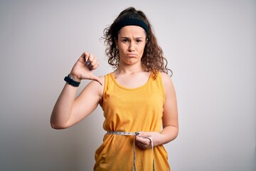 Poster - Young beautiful woman with curly hair controlling weight using tape mesure with angry face, negative sign showing dislike with thumbs down, rejection concept